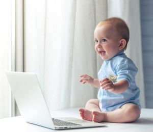 Cute little baby is looking away and laughing while sitting on desk near the laptop