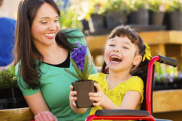 Laughing little girl in wheelchair holds lovely potted plant with a purple bloom as her smiling mom looks on