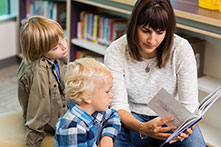 Mom pages through book in library as toddler watches intently and older child reads along beside her