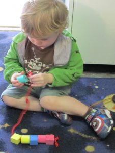 Boy toddler in a green jacket strings blocks through a red piece of string.