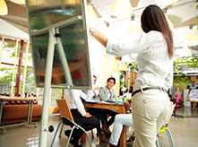 Group of people sitting around the table follow what woman is pointing out on white board stand